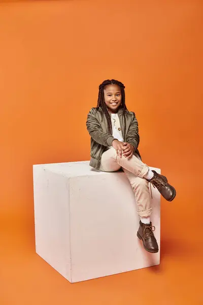 Cheerful preteen african american girl in winter attire sitting on cube and smiling at camera — Stock Photo