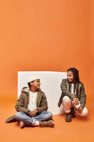 Jolly african american girl and boy in winter attires sitting near white cube smiling at each other — Stock Photo