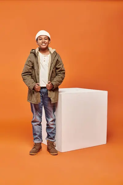 Joyeux garçon afro-américain en veste d'hiver posant à côté du cube blanc et souriant à la caméra — Photo de stock