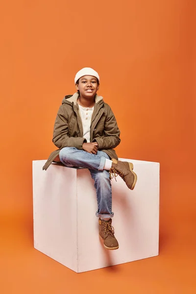 Joyeux garçon afro-américain en veste d'hiver et bonnet chapeau près de blanc cube souriant à la caméra — Photo de stock