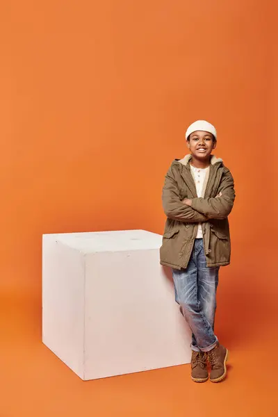 Jolly preteen african american boy in warm winter jacket posing near white cube on orange backdrop — Stock Photo