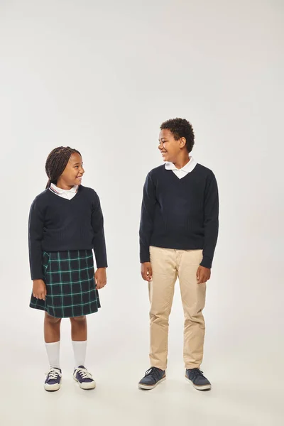 Alegres escolares afro-americanos de uniforme sorrindo um para o outro em fundo cinza — Fotografia de Stock