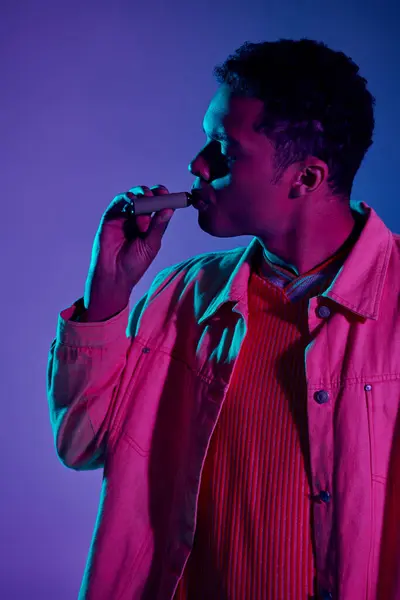Joven africano americano hombre vapeo cigarrillo electrónico en gris telón de fondo con iluminación colorida - foto de stock