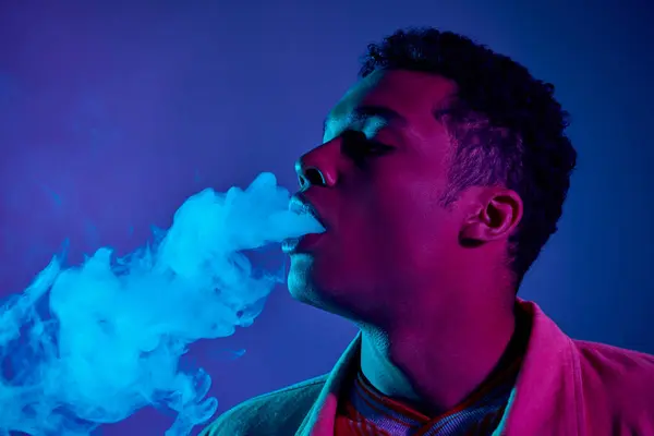 Junger afrikanisch-amerikanischer Mann atmet Rauch vor blauem Hintergrund mit violettem Licht ein — Stockfoto