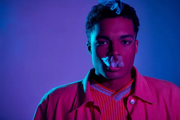 Joven afroamericano hombre exhalando humo sobre un fondo azul con iluminación púrpura, gen z - foto de stock