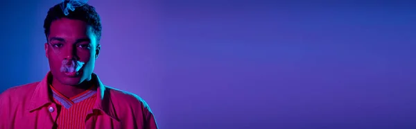 Jeune homme afro-américain exhalant de la fumée sur un fond bleu avec un éclairage violet, bannière — Photo de stock