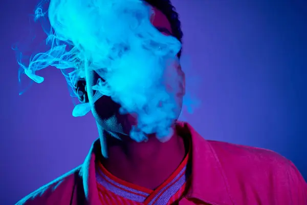 Guapo afroamericano hombre exhalando humo contra fondo azul con iluminación púrpura, gen z - foto de stock