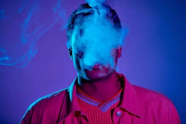 Retrato de homem afro-americano exalando fumaça contra fundo azul com iluminação roxa, gen z — Fotografia de Stock