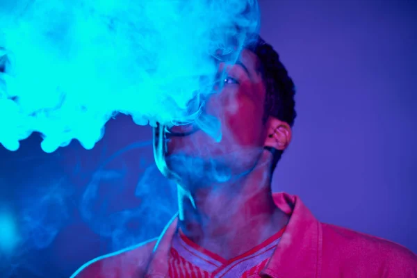 Retrato de tipo afroamericano exhalando humo contra fondo azul con iluminación púrpura, gen z - foto de stock
