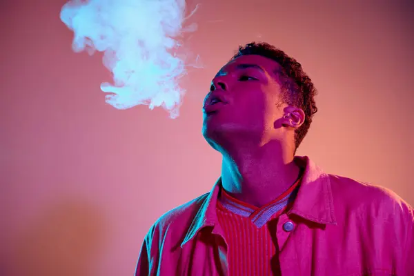 Retrato de homem afro-americano exalando fumaça contra fundo vibrante com iluminação azul, vape — Fotografia de Stock