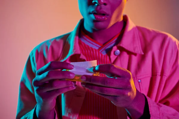 Cropped african american man rolling cigarette on vibrant background with blue neon lighting — Stock Photo