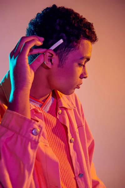 Cool homme afro-américain tenant roulé cigarette derrière son oreille sur rose pastel avec lumière bleue — Photo de stock