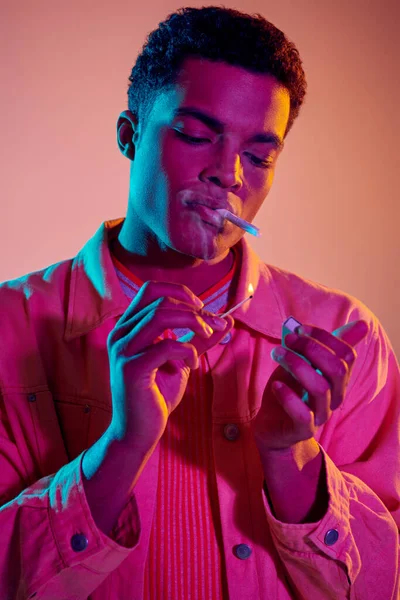 Hombre afroamericano encendiendo un cigarrillo con fósforo bajo luces de neón azul sobre fondo rosa - foto de stock
