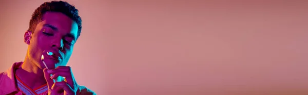 Африканский американец закуривает сигарету со спичкой под синим неоновым светом на розовом фоне, баннер — стоковое фото