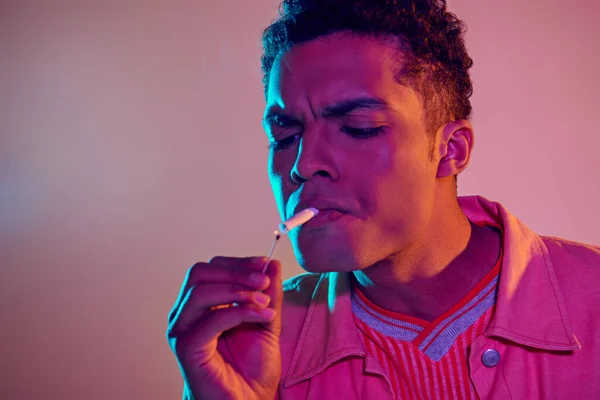Focalizzato uomo africano americano illuminazione sigaretta con fiammifero sotto luci al neon blu su sfondo rosa — Foto stock