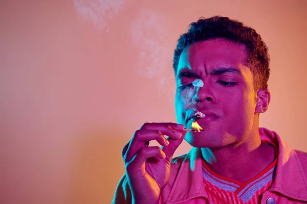 Junger schwarzer Mann zündet Zigarette mit Streichholz unter blauem Neonlicht auf rosa Hintergrund an — Stockfoto