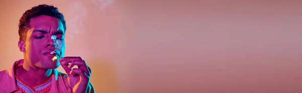 Giovane nero accensione sigaretta con fiammifero sotto luci al neon blu su sfondo rosa, banner — Foto stock