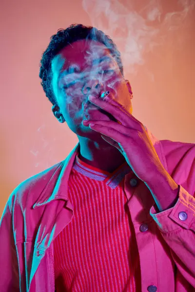Fresco y joven afroamericano hombre fumando cigarrillo bajo luces de neón azul sobre fondo rosa - foto de stock