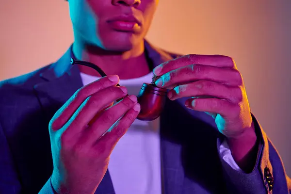 Cultivé afro-américain gars inspecter une pipe à fumer sous la lumière bleue sur fond coloré — Photo de stock