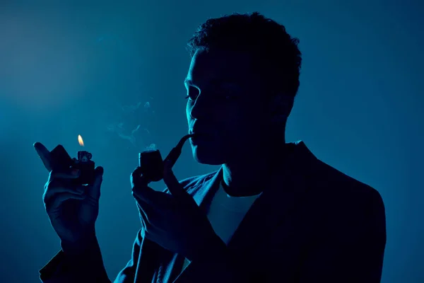 Joven afroamericano hombre sosteniendo más ligero y fumar pipa sobre fondo azul oscuro con iluminación - foto de stock