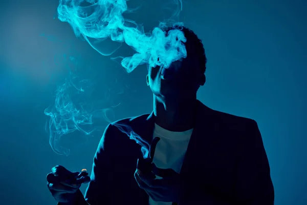 Encaracolado afro-americano homem segurando mais leve e fumar cachimbo enquanto exalando fumaça no azul escuro — Fotografia de Stock