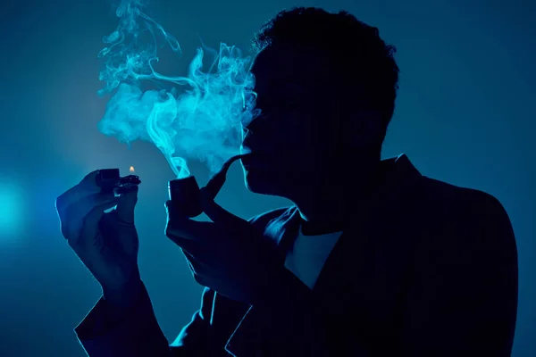 Hombre afroamericano sosteniendo el encendedor y la pipa mientras exhala humo sobre fondo azul oscuro - foto de stock