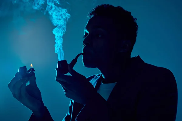 Jovem afro-americano cara segurando isqueiro e tubo enquanto exalando fumaça no fundo azul escuro — Fotografia de Stock