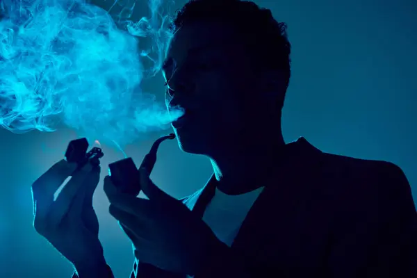Afro americano cara segurando isqueiro e tubo enquanto exalando fumaça no fundo azul escuro — Fotografia de Stock