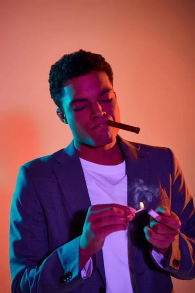 African american guy with cigar holding match with fire on pink background with blue lighting — Stock Photo
