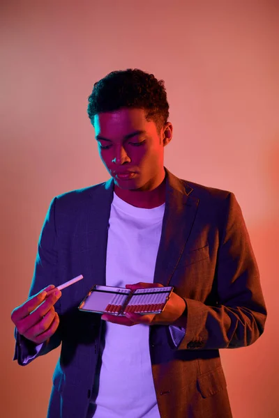 Portrait of young african american man holding cigarette case on pink background with blue lighting — Stock Photo