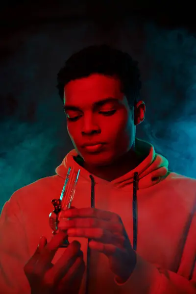 Young african american man in hoodie looking at glass bong on dark background with red lighting — Stock Photo