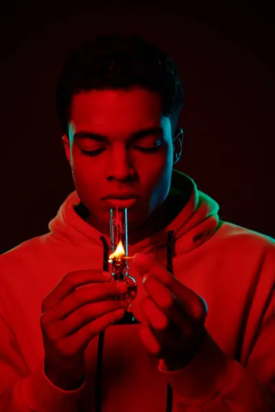 Junger afrikanisch-amerikanischer Mann in Kapuzenpullover beleuchtet Glasbong auf dunklem Hintergrund mit roter Beleuchtung — Stockfoto