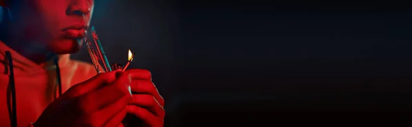 Cropped banner of black man in hoodie lighting glass bong on dark background with red lighting — Stock Photo