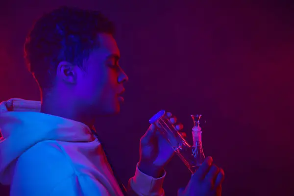 Молодой африканский американец в толстовке смотрит на стеклянный бонг на темно-фиолетовом фоне с освещением — стоковое фото