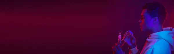 Молодой африканский американец в толстовке смотрит на стеклянный бонг на темно-фиолетовом фоне, баннер — стоковое фото
