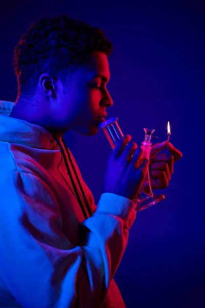 Seitenansicht, afrikanisch-amerikanischer Mann in Kapuzenpullover beleuchtet Glasbong auf dunkelblauem Hintergrund mit rotem Licht — Stockfoto