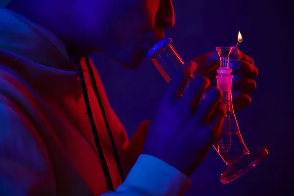 Recadré homme afro-américain à capuche éclairage verre bong sur fond bleu foncé avec lumière rouge — Photo de stock