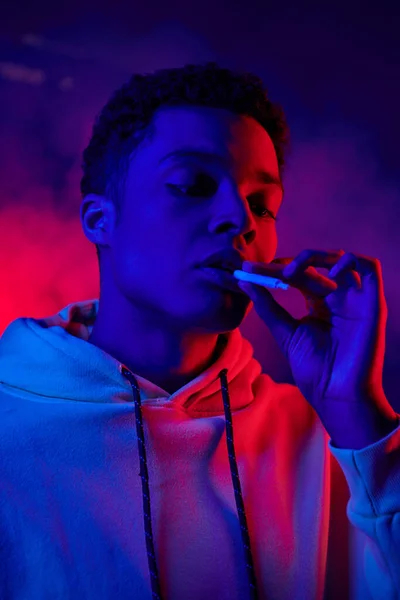 Afrikanisch-amerikanischer Mann hält Zigarette in der Hand, während er auf dunkelblauem Hintergrund mit rotem Licht steht — Stockfoto