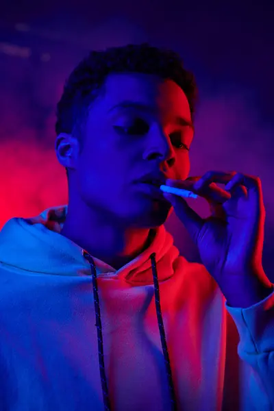 African american man holding rolled cigarette while standing on dark blue background with red light — Stock Photo