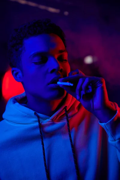 Homme afro-américain tenant roulé cigarette près de la bouche sur fond bleu foncé avec lumière rouge — Photo de stock