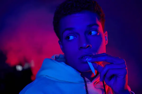 Hombre afroamericano fresco sosteniendo el cigarrillo enrollado cerca de la boca sobre fondo azul oscuro con luz roja - foto de stock