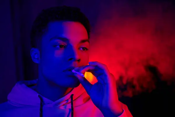 Cool homme afro-américain tenant roulé cigarette près de la bouche sur bleu foncé avec lumière rouge, fumeur — Photo de stock