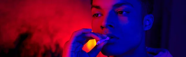 Afrikanisch-amerikanischer Mann hält Zigarette in Mundnähe auf dunkelblauem mit rotem Licht, Raucher-Banner — Stockfoto