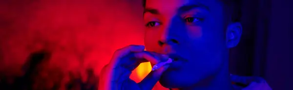 African american man holding rolled cigarette near mouth on dark blue with red light, smoker banner — Stock Photo