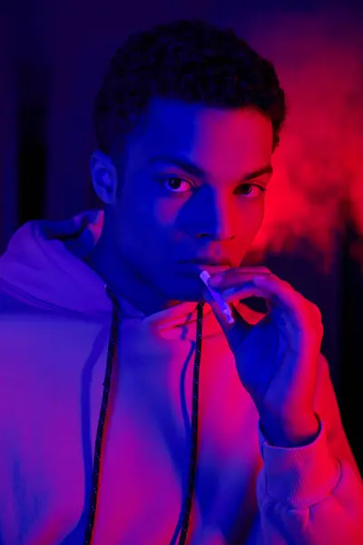 Serious african american man holding rolled cigarette on dark blue background with red light — Stock Photo