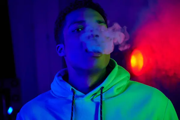 Serious african american man exhaling smoke on dark blue background with red light — Stock Photo