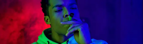 Estandarte de afroamericano joven en sudadera con capucha fumar cigarro sobre fondo azul oscuro con luz roja - foto de stock