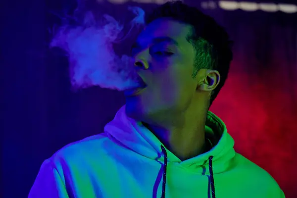 Homme afro-américain grave expirant fumée sur fond sombre avec lumière bleue et rouge, fumeur — Photo de stock