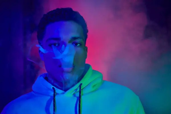 Cool african american man exhaling smoke on dark background with blue and red light, smoker — Stock Photo