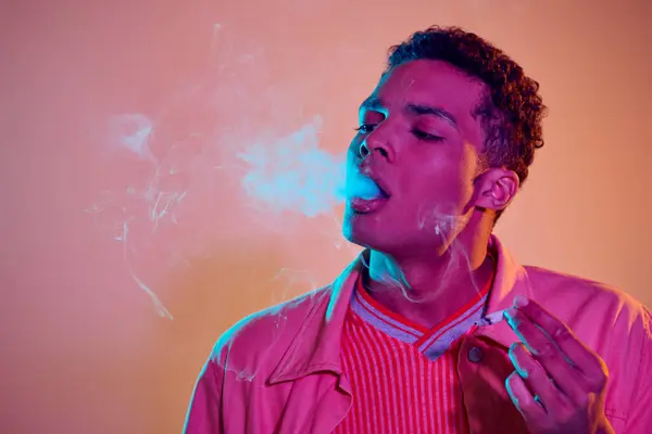 Cool african american man exhaling smoke on bright background with neon light, smoker with cigarette — Stock Photo